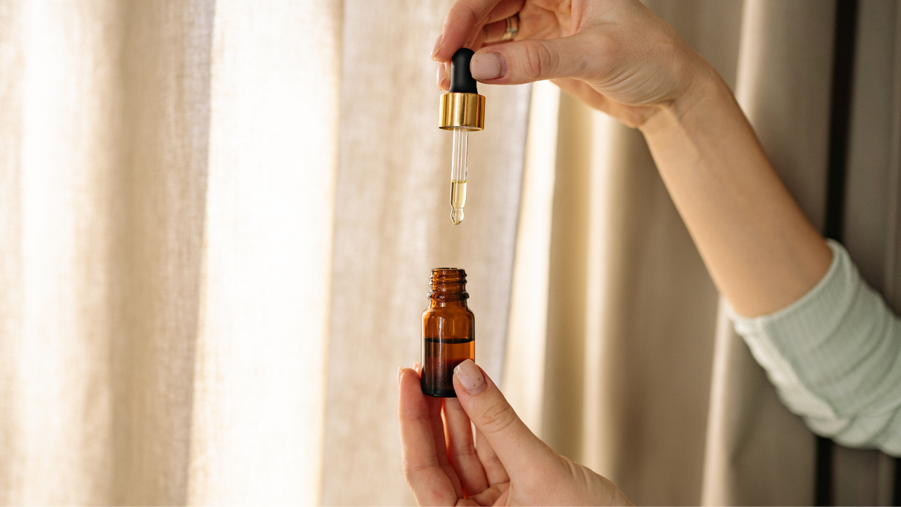 Hand holding a small amber glass dropper bottle with a gold-trimmed pipette dispensing essential oil.