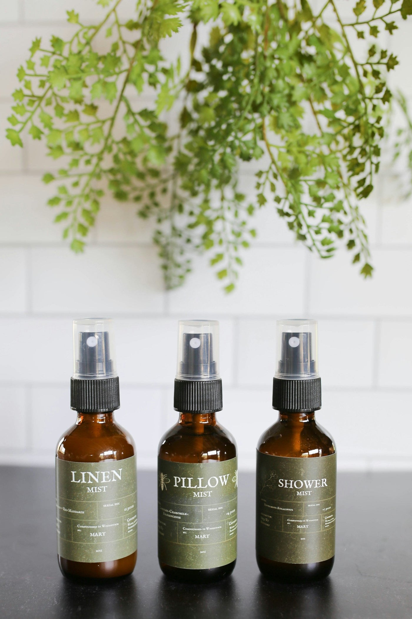 Three amber glass spray bottles with black premium mist closures against a white tiled background.