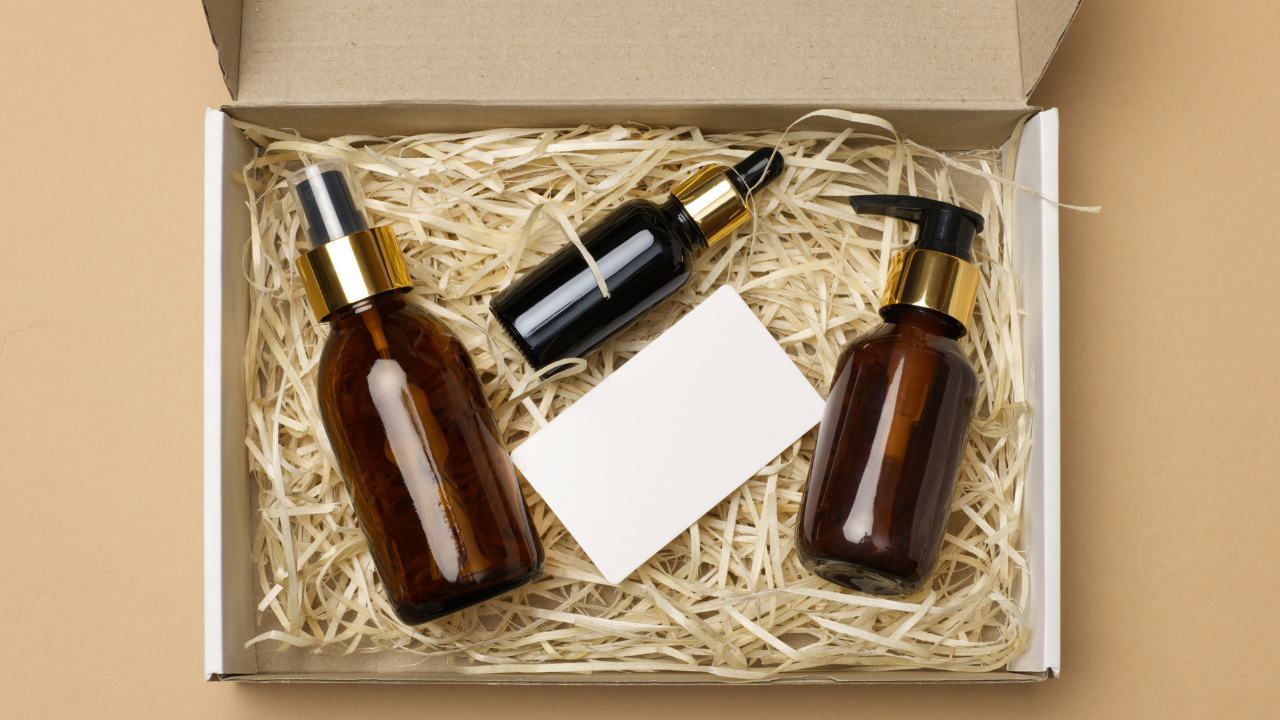 Amber glass cosmetic bottles with gold pumps and a blank white label inside a cardboard box with wood wool filler.