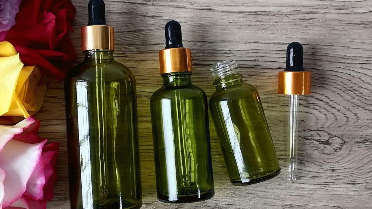 Three green glass essential oil bottles with gold dropper caps arranged on a wooden surface next to colorful roses.