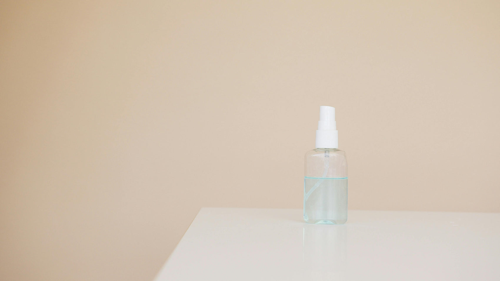 Clear glass spray bottle filled with liquid sitting on a white surface against a neutral beige background.