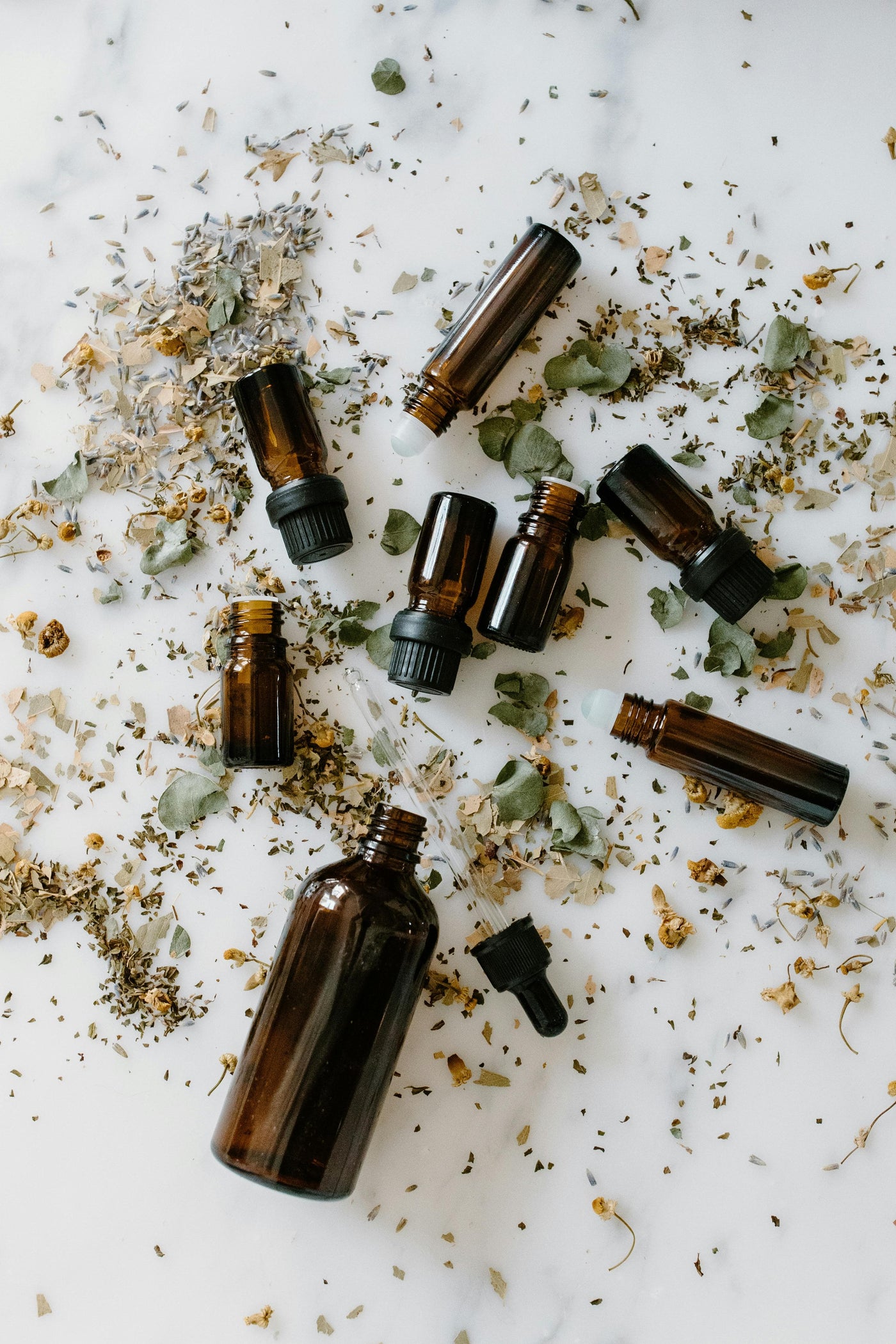 Amber glass dropper and roller bottles scattered on a white marble surface with dried herbs and flower petals.