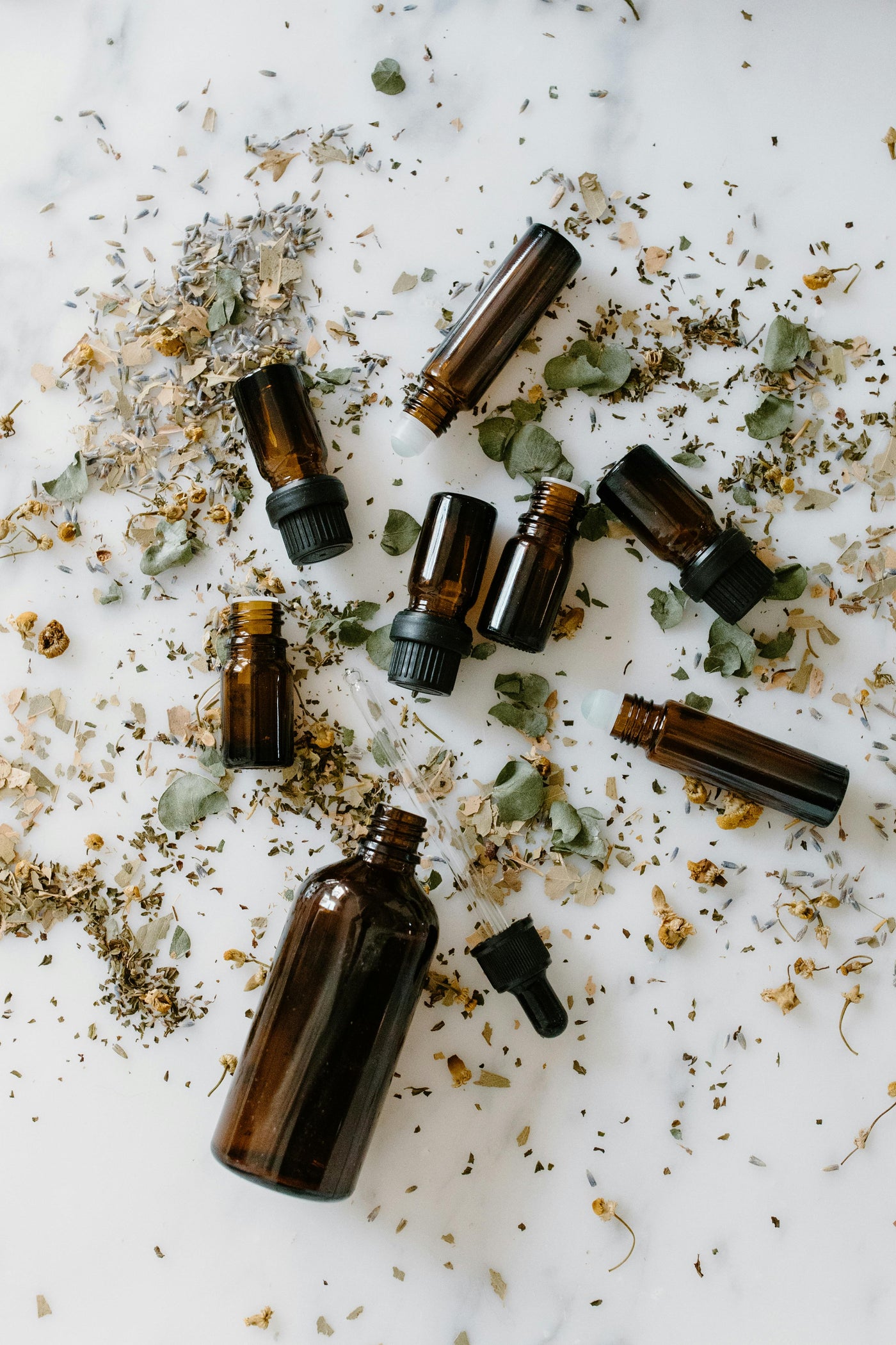 Amber glass dropper bottles and roller vials scattered on a white marble surface with dried herbs and flowers.