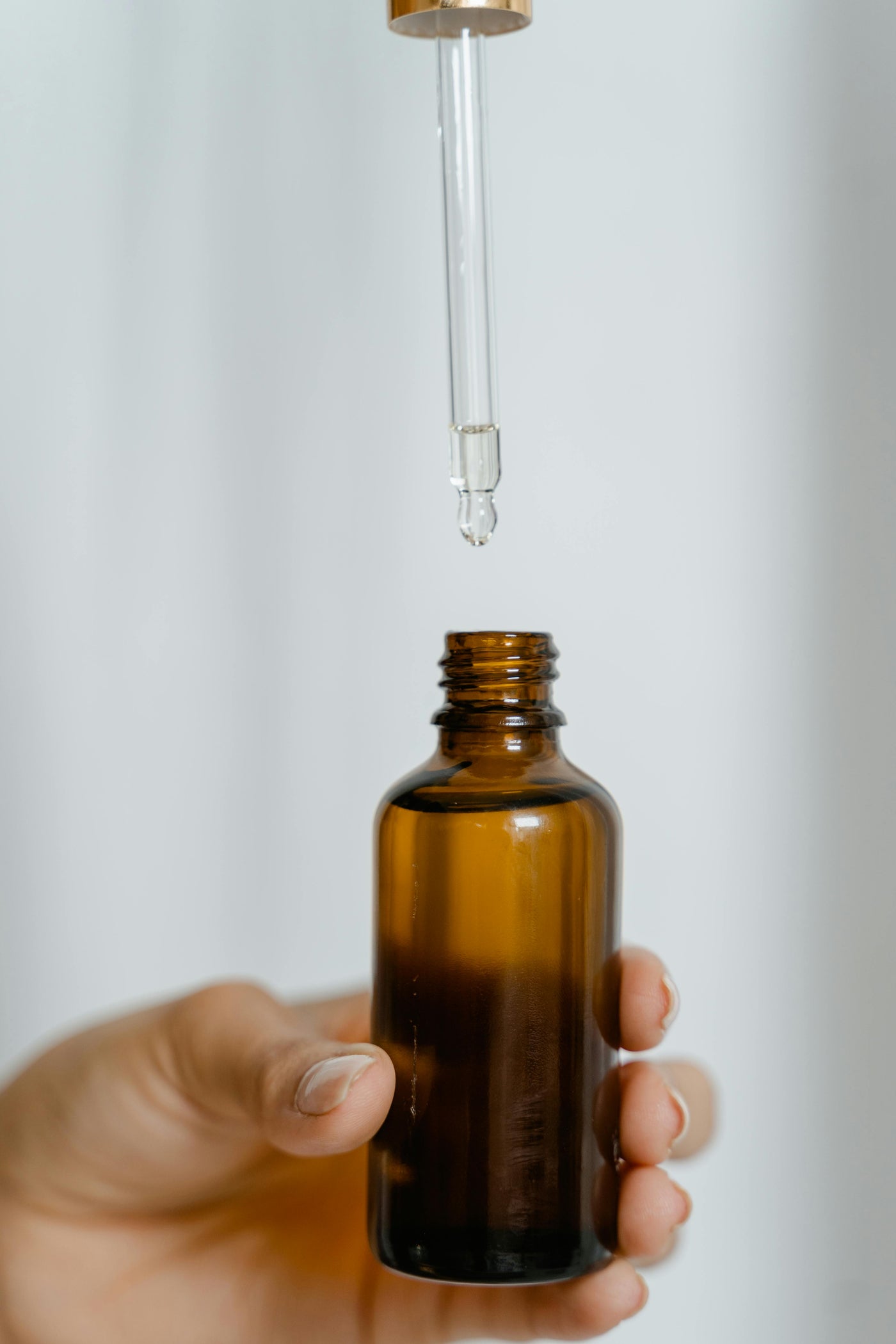 Hand holding an amber glass dropper bottle with a clear serum drop falling into the opening.
