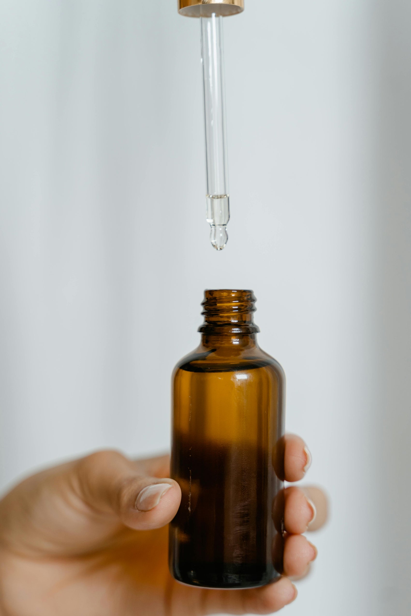 Hand holding an amber glass dropper bottle with a clear serum drop falling into the opening.