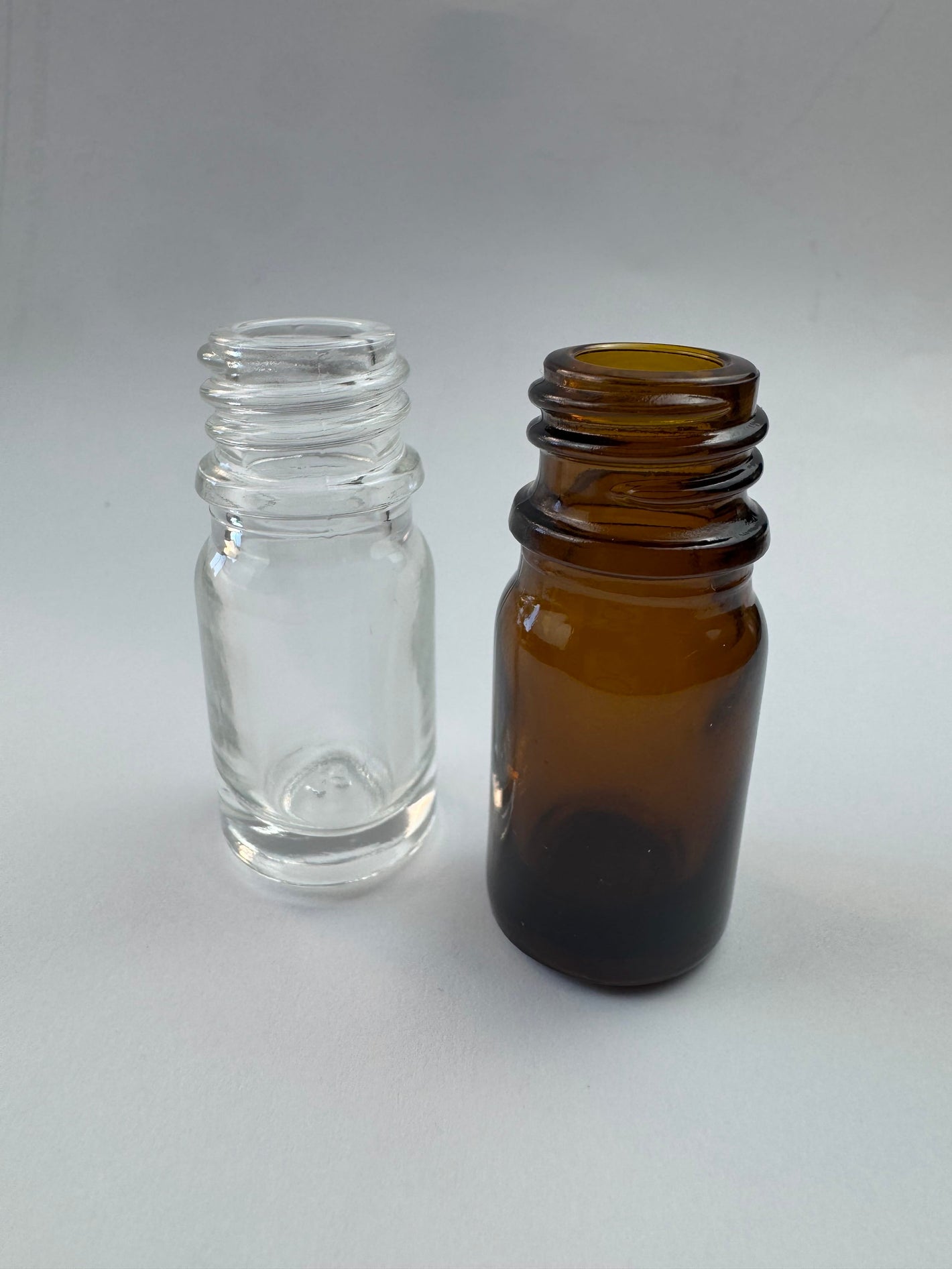 Two empty 5ml glass dropper bottles, one clear and one amber, standing on a neutral white background.