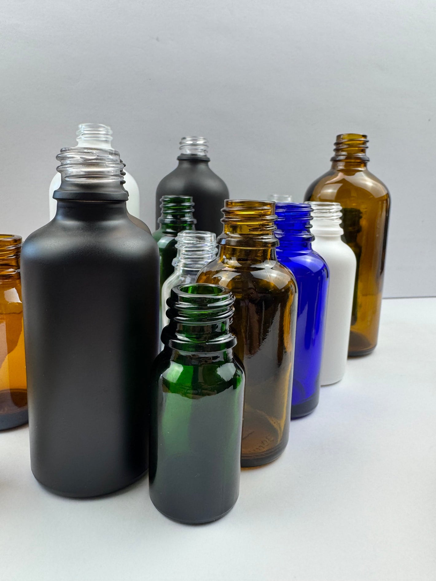 Assorted 20ml glass dropper bottles in matte black, amber, cobalt blue, green, and white finishes.